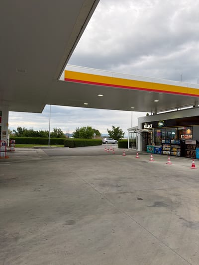The image depicts a Shell gas station in Dragor, Bulgaria. The sky is overcast. A Shell Select convenience store is visible with an assortment of snacks and beverages. A white car is parked beyond the store. Orange and white cones are placed near the store entrance. Green hedges are present in the background. The concrete ground appears to be stained.