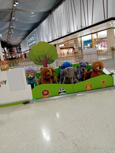 The image shows an indoor mall scene in Amadora, Portugal, with a row of stuffed animal ride-on toys visible. The animals include a lion, koala, elephant, bear, and other characters, arranged in a line as if sitting in a vehicle with bright green sides adorned with animal print images. The ride is decorated with a green tree attached to the side. The ride appears to be an attraction for children, set in a brightly lit mall with retail stores visible in the background. The floor is a light speckled colour, likely tiled.