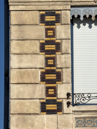 The image shows a close-up of a building facade in Charnay-lès-Mâcon, France. The facade is constructed from large, light-colored stone blocks, horizontally layered. A vertical decorative element made of alternating dark brown and yellow bricks is embedded within the stone. This brickwork forms an elongated, symmetrical pattern with wider sections at the top and bottom and a narrower middle. To the right of the brickwork is a window with a white roller shutter. Above the shutter, there is a decorative metal grille with intricate scrollwork and leaf motifs. Below the window, there is a section of ornate blue tiles with floral patterns. The overall architectural style appears to be a blend of traditional stone construction with decorative brick and metal elements.