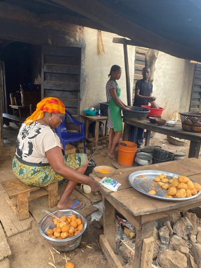 Three people are outside near a building, possibly cooking. A woman in a patterned skirt and headscarf sits on a low stool with a basket of round fried food and tongs. A tray of similar food sits nearby. Behind her, a woman in a green apron holds a black bucket. Further back, a man in a black shirt also holds a black bucket. Cooking utensils and pots are scattered on a table near what appears to be a fire.