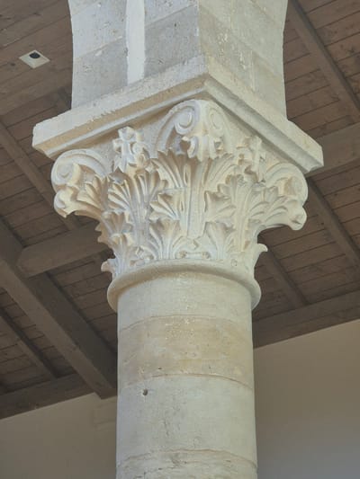 The image shows a close-up of a stone pillar with ornate carving on its capital. The pillar is part of the architecture of the Crusader Monastery of Abu Gosh, located in Abu Gosh, Israel. The capital is decorated with acanthus leaves and volutes, a style characteristic of Romanesque architecture. Above the pillar, wooden beams and ceiling planks are visible, along with a square light fixture embedded in the ceiling. The pillar itself is made of roughly dressed stone blocks. The overall scene suggests an interior or covered area within the historic monastery.