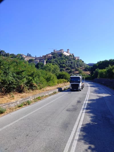 A white Volvo truck drives on a paved road in a rural area under a clear blue sky. The truck has a flatbed trailer and is positioned in the right lane of a two-lane road. The road curves to the right, with a stone wall on the right side and a vegetated embankment on the left. In the background, a fortified town is situated on a hill, with several buildings and a crane visible. The town appears to be medieval or historic in architecture. The environment is characterized by lush green trees and dry, brown grass on the embankment. The sunlight suggests it is daytime and warm. The image is taken from a slightly elevated perspective, likely from another vehicle. The location is Spoleto, Italy, and the fortified town in the background is likely the Rocca Albornoziana and the historic center of Spoleto.