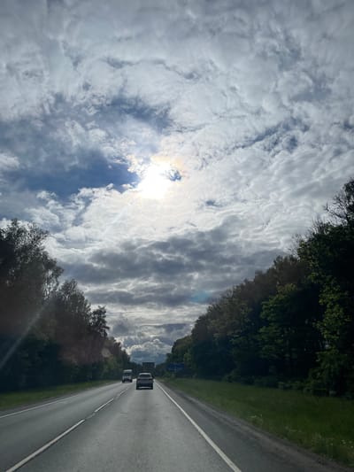 The image shows a view from a vehicle driving on a highway, potentially near Ol'khovka, Russia, but the specific location cannot be confirmed. The sky is filled with predominantly white and grey clouds, with the sun partially visible through a break in the cloud cover. The roadway is asphalt and has a white lane divider. Two vehicles are visible ahead, seemingly traveling in the same direction. The roadside is lined with trees. The image is oriented with the sky on the left and the road on the right.