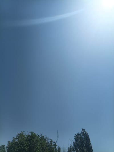 The image depicts a clear, bright blue sky with a prominent sun flare originating from the upper right quadrant. The lower portion of the image features the tops of several trees with dense green foliage, characteristic of a temperate climate. The scene appears to be an outdoor environment. Given the information available, it is not possible to pinpoint the exact location within the city of Нуъмон Розиқ, Tajikistan, but the imagery suggests a natural or park-like setting within or on the outskirts of an urban area. There are no people or specific objects visible that would indicate a particular activity or event.