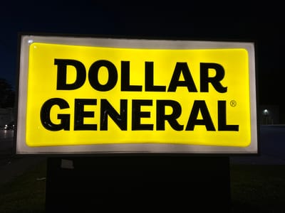 This is a photograph of a backlit Dollar General sign at night. The sign is rectangular with rounded corners and is illuminated with a bright yellow light. The words "DOLLAR GENERAL" are written in large, black, bold, sans-serif font across the sign. The registered trademark symbol "®" is present to the right of "GENERAL". The sign is mounted on a black pole structure. In the background, the sky is dark blue, indicating nighttime. To the left of the sign, faint lights from a street and distant buildings are visible. The area below the sign appears to be a grassy area with some litter. The overall setting suggests a commercial area in a city.