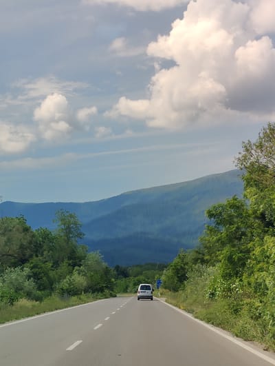 A white van travels down a two-lane asphalt road surrounded by lush green trees and vegetation. In the distance, a series of blue-hued mountains rise against a sky filled with dramatic, fluffy white clouds. The road has white dashed lines indicating lanes and a solid white line along the right edge. A blue road sign is visible on the right side of the road, near the van. The scene appears to be a scenic drive, possibly in the vicinity of Mezdreya, Bulgaria, given the mountainous terrain.