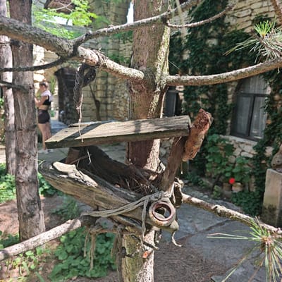 The image depicts an outdoor scene in Останинское сельское поселение, Ukraine, dominated by a rustic, handmade structure, likely a bird feeder, constructed from weathered wooden planks and branches. This structure is secured to the trunk of a tree, possibly a pine, with thick, coarse rope, and includes a rusty metal ring. Pine needles and debris are visible within the structure. To the left, another tree trunk stands.
In the mid-ground, a person, blurred due to depth of field, is partially visible to the left of the central tree. They appear to be wearing a dark top and shorts, with a light-colored cloth draped over one shoulder. Behind the trees, a building with textured stone walls is prominent, featuring an arched entryway and dark-framed windows. Portions of the building's facade are covered in dense green ivy. The ground is a mix of bare earth, scattered pine needles, and irregular stone paving. A single red flower is visible on the ground near the base of the stone building on the right. The scene is brightly lit by natural sunlight, casting shadows and highlighting textures.