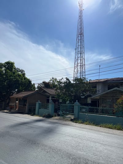 The image depicts a rural street scene in Bukitrata, Indonesia, under a bright, partly cloudy sky, suggesting it is daytime. A prominent subject is a tall, lattice-style telecommunications tower dominating the left side of the frame, its metal structure silhouetted against the sun. A network of power lines stretches across the upper portion of the image, originating from the tower and extending towards unseen poles.

To the right of the tower, a row of houses with tiled roofs lines the street. The architecture appears modest, with some houses featuring decorative metal fencing and greenery, including trees and grass, growing along the property lines. The road itself is paved and appears to have some tire marks. The scene is quiet and appears to be a residential area. No people are visible in the frame, and no text is present. The overall impression is one of a typical day in a developing area with essential infrastructure present.