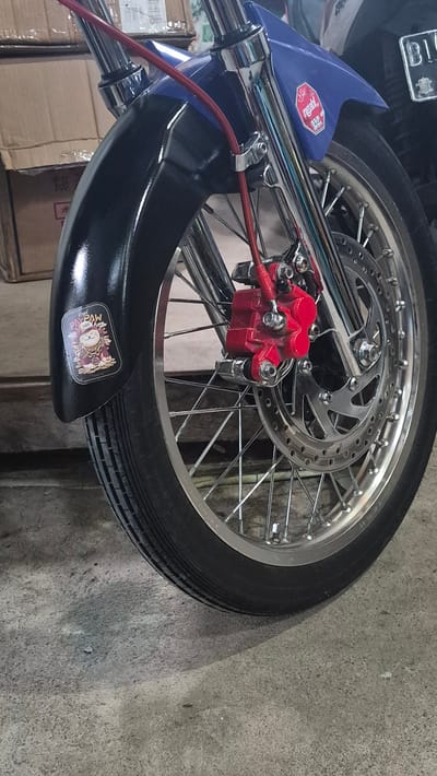 The image shows a motorcycle front wheel with a disc brake and a red brake caliper. The wheel has chrome spokes and rim. The tire is black. The front fender is black with a sticker of a cat-like creature. The brake line is red. Part of the motorcycle's front suspension is visible, with blue and chrome elements. The scene is set indoors, on a concrete floor. Boxes are visible in the background. The location is Pante Aree, Indonesia, according to EXIF data.