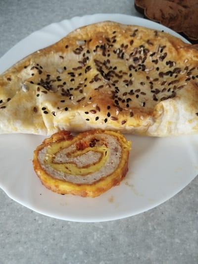 A white plate holds a baked item resembling a flatbread topped with various seeds, including black sesame, white sesame, and flax seeds. Adjacent to it is a slice of rolled food, showcasing layers of meat and what appears to be an egg-based filling, with an orange exterior. The plate sits on a speckled gray surface, likely a countertop. The background is out of focus, with a glimpse of a brown object on the right. There are no people or animals visible in the image. The overall scene suggests a meal or a food presentation. The image was taken in Bezlesnyy, Russia.