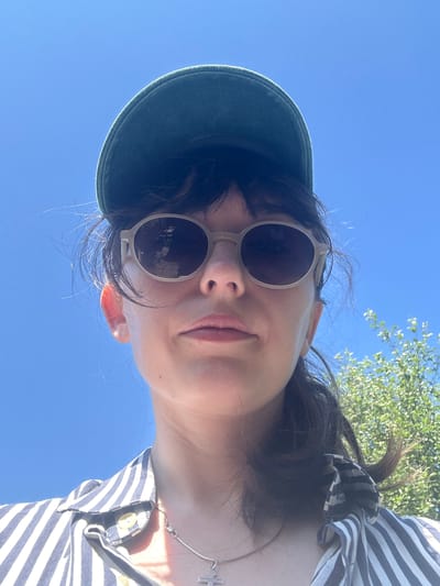 This is a close-up, low-angle selfie of a young woman outdoors, likely on a sunny day. She is wearing a dark green baseball cap and light-colored round sunglasses. Her hair is dark and appears somewhat windswept. She is also wearing a striped shirt with buttons and a necklace with a cross pendant. The background consists of a clear blue sky and the green foliage of a tree. The woman's expression is neutral, with her lips slightly parted. The image is shot from a bottom-up perspective, with the sky taking up a significant portion of the frame. There is no visible text in the image.