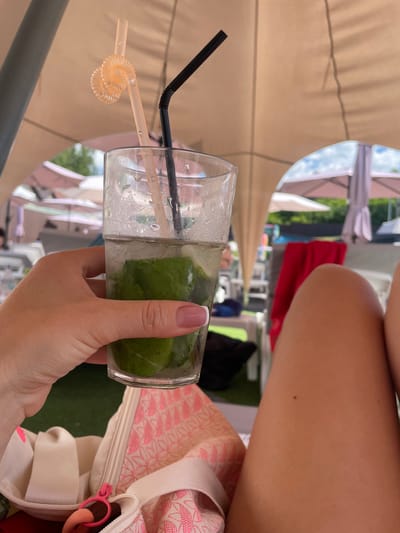An overhead perspective shows a person's hand holding a clear glass filled with a beverage and ice, with several sprigs of mint visible. Two straws are inserted into the glass: one is black and the other is clear and spiraled. The hand has manicured nails painted with a French manicure. In the background, a light-colored canopy shades the area, with glimpses of umbrellas and lounge chairs outside. A patterned bag is visible in the lower left corner. The scene suggests a relaxed outdoor setting, possibly a cafe or poolside area in Zazymia, Ukraine.