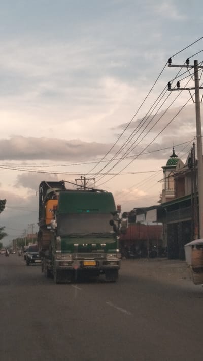 This image captures a street scene in Dua Paya, Indonesia, likely during the late afternoon or early evening, judging by the soft lighting and the presence of clouds. The primary focus is a large, green truck carrying a yellow construction vehicle on its trailer, which is in motion on the asphalt road. The truck's headlights are on, indicating dimming light conditions. To the right of the truck, a building with a distinctive green-domed structure is visible, suggesting a place of worship or a significant landmark. A utility pole with multiple wires runs across the upper portion of the frame, and other vehicles, including a black car, are present on the road, indicating ongoing traffic. The environment appears to be urban or semi-urban, with buildings lining the street and electrical infrastructure overhead. The overall impression is one of daily life and activity in an Indonesian town.
