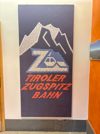 This image shows a close-up view of a framed tapestry-like artwork depicting mountains and the words "Tiroler Zugspitz Bahn" in red text. The artwork is hung on a pale pink wall next to a dark grey baseboard. To the right, a section of bright orange wall is visible. Above and to the right of the main artwork, a smaller framed photograph of a mountain landscape is partially visible. The artwork itself has a textured appearance, possibly indicating it is made of embroidered or woven material, with white and grey stitching for the mountains, blue and white for a stylized "Z" emblem with a cable car, and red stitching for the text. The overall scene appears to be indoors, likely in a public space such as a tourist information center or a transport hub in Ehrwald, Austria, given the reference to the Zugspitz Bahn, a cable car operating from Ehrwald to the Zugspitze mountain. There are no people or actions depicted in the image.
