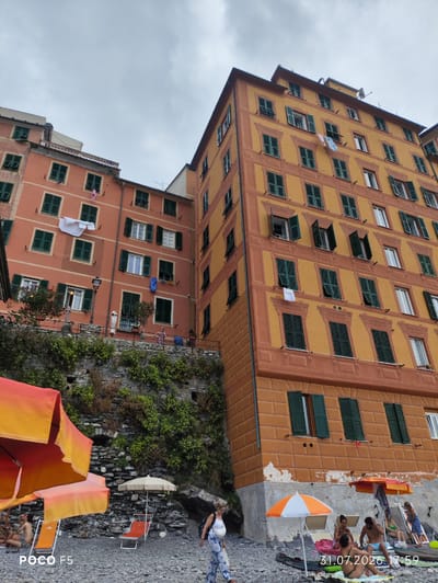 This image depicts a view of the colorful buildings of Camogli, Italy, from a beach. The buildings are tall and narrow, painted in shades of orange and pink, with dark green shutters on the windows. Laundry is hanging from several windows, adding a lived-in feel to the scene. The beach itself is made of pebbles, and several beach umbrellas and chairs are set up. In the foreground, a woman is walking on the beach, carrying a backpack. Other people are visible in the background, relaxing on the beach. The sky is overcast, suggesting it may be a cloudy day. The overall impression is one of a charming coastal town on a slightly subdued day.