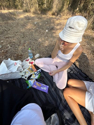 Three individuals are gathered on a dark, patterned blanket laid over dry, sparse grass and scattered rocks. The background consists of natural vegetation, including trees and shrubs, consistent with an uncultivated outdoor area in the Modi'in-Maccabim-Re'ut region of Israel.

One individual, positioned centrally, wears a white tank top, light pink long pants, and a white bucket hat bearing the "JACQUEMUS" brand name; their right hand is raised near their face. To the left of this individual, a white canvas tote bag labeled "Athletics" is visible, along with a pair of white sneakers featuring pink interiors. A plastic water bottle with a green cap is also present. Several snack items are arranged on the blanket, including bags of chips (one with "Minions" branding), a bag of "Shugi" branded snacks, and two purple "Milka Grain" chocolate bars. Portions of two other individuals are visible: one at the bottom-left, wearing black trousers and a white top, and another at the bottom-right, showing tanned legs, indicative of wearing white shorts or a skirt. The scene is illuminated by natural light.