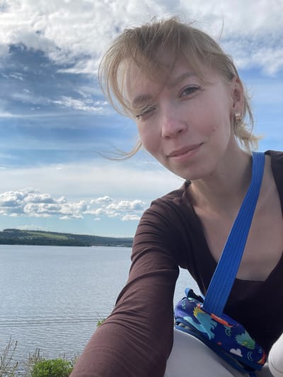 A young woman is captured in a low-angle selfie, looking upwards at the camera with a neutral to slightly smiling expression. She has blonde hair, with some strands appearing windswept, and light-colored eyes. She is wearing a form-fitting, dark brown long-sleeved top. A bright blue strap extends across her chest, to which a small blue pouch or bag is attached. This pouch features colorful, cartoon-like illustrations of dinosaurs and clouds.

The setting is outdoors under a bright, partly cloudy blue sky, with scattered white cumulus clouds. Behind the woman, a wide expanse of water, likely a large lake or river, stretches out, displaying subtle ripples on its surface. In the distance, a narrow strip of land with a dark green treeline is visible along the horizon. The lighting indicates it is daytime, suggesting clear and fair weather. The scene portrays a tranquil natural environment, located in Пятый километр, Russia. No visible text is present in the image.