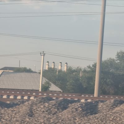 The image shows a view from a moving vehicle on a clear day in the city of Arys, Kazakhstan. In the foreground, a railway track with ballast and concrete sleepers is visible. Behind the railway, a building with a corrugated metal roof and a black roof is present. Several power lines and a utility pole are also visible. In the middle ground, trees form a dense canopy. In the background, a row of minarets, characteristic of a mosque, can be seen rising above the trees. The sky is overcast with a pale blue hue, and sunlight casts shadows on the ground. The image appears to have been taken during daytime, likely in the afternoon, given the lighting and the presence of shadows. There are no people or animals visible in the frame.