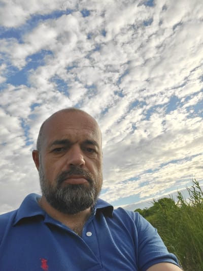 The media file is a portrait-style, selfie-style photograph taken outdoors. The primary subject is a bald, middle-aged man with a beard, wearing a blue polo shirt with a small red logo on the chest. He is looking directly at the camera with a neutral expression.
The background is dominated by a bright blue sky filled with scattered, fluffy white clouds, suggesting fair weather. To the right of the frame, there is a strip of lush green grass or foliage, indicating a natural outdoor setting, possibly a field or park. The low angle of the shot, with the sky taking up a significant portion of the image, emphasizes the outdoor environment.
The lighting suggests it is daytime, likely late morning or afternoon, given the brightness of the sky and the illumination of the subject. There are no discernible objects other than the man and the natural elements. The overall mood is casual and relaxed.
While the provided location context is "Hetmanivka, Ukraine," there are no specific visual cues within the image itself that confirm this location. There is no visible text in the media file. The image is rotated 90 degrees counter-clockwise, meaning the man's head is to the left of the frame.