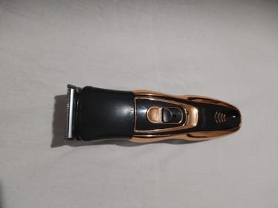 This media file shows a close-up, top-down view of a black and rose gold electric hair clipper. The clipper is lying horizontally on a textured, light-colored surface, possibly a fabric. The clipper is sleek and modern in design. The left side features the metallic shaving head with visible teeth. Moving towards the right, there's a black body section followed by a rose gold metallic section that houses a circular on/off switch with a black toggle. Further to the right, the rose gold casing continues, tapering to a point, and features the brand name "Geemy" printed in black. Below the brand name, there are three black chevron-like markings. The lighting suggests an indoor setting, possibly during the daytime, as there are no strong shadows indicating artificial light. There are no people visible in the image, and the focus is solely on the object. The overall impression is of a product shot, highlighting the details of the hair clipper. The location context provided is Moara Domnească, Romania.