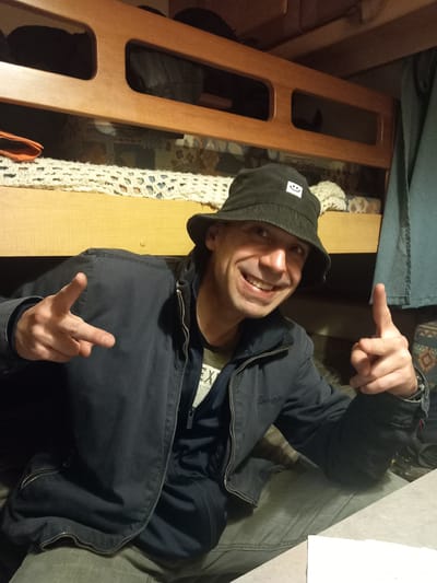 The image shows a man wearing a black bucket hat and a dark jacket, giving a peace sign with his left hand and gesturing with his right hand. He is smiling and looking towards the camera. The background appears to be the interior of a room with wooden bunk beds and bedding. There is no discernible information within the image that indicates the location as Shotton, United Kingdom.