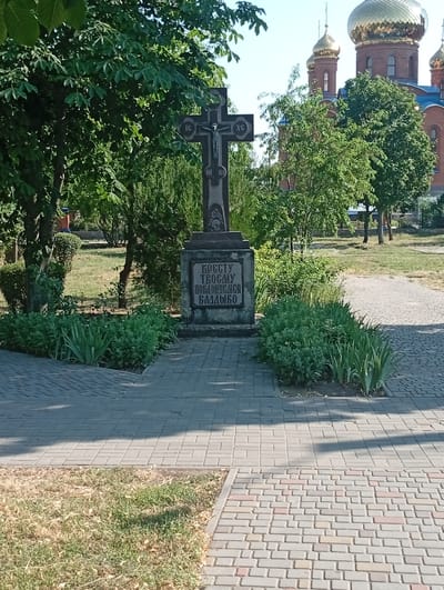A dark, stone cross stands prominently in the foreground, featuring a depiction of Jesus Christ. The cross is mounted on a stone pedestal with engraved text. Behind the cross, lush green trees and bushes create a vibrant backdrop. In the distance, a large, red church with golden domes is visible, suggesting a religious setting. The scene is bathed in bright sunlight, indicating a clear, sunny day. The ground is paved with cobblestones, and there is a patch of dry, yellowed grass to the left of the path. The overall atmosphere is peaceful and serene. The engraved text on the pedestal appears to read: "КРЕСТУ ТВОЕМУ ПОКЛОНЯЕМСЯ ВЛАДЫКО". The image is captured from a slightly low angle, looking up towards the cross and the church. The time of day is likely daytime, given the strong sunlight and clear sky. The location is identified as Pokrov, Ukraine.