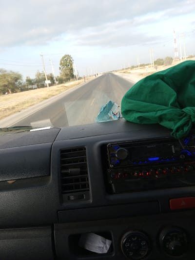 The photo shows a vehicle's dashboard, a Sony car stereo prominently displayed. A green cloth rests on the right side of the dashboard, and a small stack of paper or money is near the windshield. The view through the windshield captures a long, straight road under a cloudy sky with power lines and distant trees. The setting appears rural, with vegetation along the roadside.