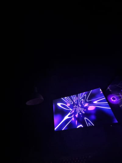 The scene depicts a dark indoor environment, potentially a nightclub or entertainment venue, in Petkovo, Bulgaria. A rectangular display screen, illuminated with vibrant purple and pink abstract patterns featuring white neon-like lines and concentric circles radiating from a central point, is positioned prominently on the right. To the left, a single, inverted, cone-shaped light fixture is partially visible, appearing to be off or unlit. A faint circular purple glow or reflection is visible on the far right edge of the frame. The overall setting is dimly lit by the display screen's luminescence. No individuals are visible.