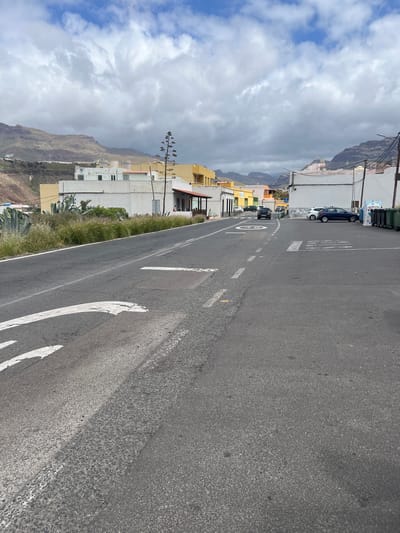 The photograph depicts a street scene in the town of La Aldea de San Nicolás, Spain. The primary focus is the asphalt road stretching into the distance. Road markings, including a dashed white line dividing lanes, a painted speed limit of "30", and the word "STOP" painted on the road surface, are visible.

Buildings line the left side of the road. Some appear to be residential, with white and yellow painted walls. Mountains rise in the background, partially obscured by cloudy skies.

Several parked cars are on the left side of the road. The sky is filled with a mix of white and gray clouds. Vegetation, including trees and shrubs, grows along the roadside.
