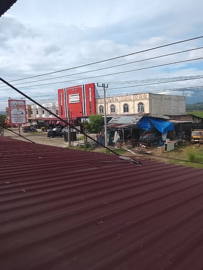 The image depicts a street scene in Manyang Cut, Indonesia, with several buildings and vehicles visible. In the foreground, a dark red, corrugated metal roof dominates the lower portion of the frame. Behind the roof, a street runs horizontally, with a line of buildings on the right side and a wider road with some parked cars and a motorbike on the left.

The building on the far left has a large billboard with a picture of several people. Adjacent to it is a white building with a red and white facade, featuring prominent text that reads "DEWAN PENGURUS WILAYAH (DPW) PIDIE JAYA". To the right of this building is a cream-colored structure with arched windows. Further to the right, a makeshift structure with a corrugated metal roof covered partially by a blue tarp is visible. Several vehicles are parked or stored under this structure, including a dark-colored SUV and a yellow truck. The background shows a hilly landscape with green fields under a cloudy sky. Several electrical wires stretch across the upper part of the image. There are no people clearly visible in the frame, only vehicles and buildings. The scene appears to be a commercial or administrative area.