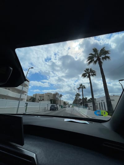 The image depicts a street scene in Sant Llorenç des Cardassar, Spain, viewed from the interior of a dark-colored vehicle through its windshield. The time of day is daytime, under a partly cloudy sky with bright sunlight.
The foreground is dominated by the vehicle's dark dashboard and the upper interior roofline. On the left side of the dashboard, a dark infotainment screen is partially visible. A piece of paper or map rests on the dashboard in the center. A circular blue and yellow sticker, consistent with a Spanish DGT environmental label, is affixed to the lower right corner of the windshield, displaying partial numbers "00" and "041".
The street extends into the distance, appearing as a paved road with vehicles. On the left side of the road, a multi-story white building with curved balconies is prominent. Further along this side, several green electric vehicle charging stations are visible, followed by a dark-colored SUV parked at the curb, facing away from the viewer. Other buildings are visible beyond.
On the right side of the road, a prominent line of tall palm trees borders the street. Behind them, white low-rise buildings are visible. A pedestrian wearing a high-visibility yellow/green vest is walking away from the camera on the sidewalk or shoulder, positioned midway down the right side of the street. Multiple streetlights are visible on both sides of the road.
