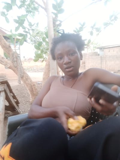 The image depicts a person seated outdoors, holding a yellow object in one hand and a dark-colored mobile phone in the other. The individual wears a tan top and dark-colored pants. The background includes a tree with green foliage and a building in the distance, suggesting an urban or semi-urban environment. The location is identified as Uke, Nigeria, based on user prompt.