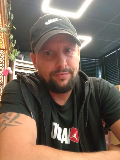 A man with a dark beard and mustache, wearing a black Nike cap, a black Jordan brand t-shirt under a black zip-up hoodie, is looking at the camera. A tattoo is visible on his left arm. In the background, there is a decorative screen with green plants, a square lighting fixture on the ceiling, and blinds. The location is Cenon, France.