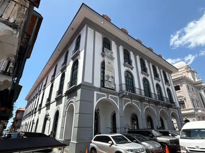 The image displays the Museo del Canal Interoceánico de Panamá, situated in the Casco Antiguo of Panama City, Panama, facing the Plaza de la Independencia. The primary subject is a multi-story building with a white facade and gray architectural accents, including arched ground-floor porticoes and vertical pilasters. Numerous arched windows on the upper floors are fitted with black metal balconies. A large banner on the building's facade explicitly identifies it as the "MUSEO DEL CANAL INTEROCEÁNICO DE PANAMÁ." To the left, a portion of an older, distressed building with wooden balconies is visible. To the right, a grand, light-pink building featuring elaborate architectural details and a dome is partially visible. The scene is illuminated by natural daylight under a clear blue sky with scattered white clouds. The street in the foreground contains several parked vehicles, including a silver SUV, a dark-colored sedan or SUV, a silver pickup truck, and a white van. An orange traffic cone is visible near the parked cars. A black vehicle with a dark canopy is partially present in the lower-left corner. No individuals are clearly discernible.