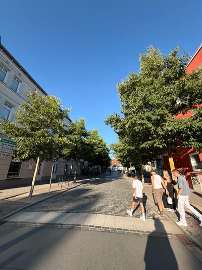 This image captures a sunny day scene on a cobblestone street in Rostock, Germany, likely in the Altstadt (Old Town) area, given the architectural style and narrow street. A group of four people are walking across the street.
From left to right:
1. A young person in a white t-shirt and dark shorts is walking, casting a long shadow.
2. A person in a white tank top and patterned shorts is walking.
3. A person in a black dress is walking.
4. A person in a grey t-shirt and white pants is walking.
They appear to be moving away from the viewer, towards the right side of the image. The street is lined with trees, which are leafy and green, indicating a warm season. On the left side of the street, there is a light-colored building with windows and what appears to be signage indicating directions such as "Toilette", "Kleine Komödie", and "Polizei". On the right side, a building painted in a vibrant red hue stands out, with windows and graffiti visible on its lower section. The sky is a clear, bright blue. The sun is casting strong shadows, indicating it is likely mid-afternoon. In the background, more trees and buildings are visible, suggesting a well-established urban environment.