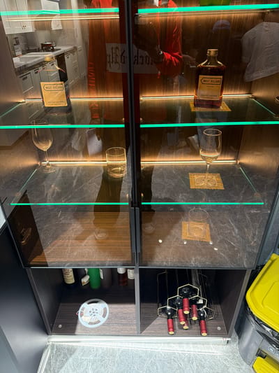 The image displays a modern, illuminated display cabinet, appearing to be rotated 90 degrees clockwise from its natural upright position. The cabinet features multiple glass shelves, framed by integrated LED lighting strips that emit both warm amber and greenish hues, creating a sophisticated ambiance. The structure combines dark wood or laminate panels with a marble-like pattern.
On the upper left shelf, an amber-colored glass bottle is prominently displayed, featuring a label that reads "WEST VILLAGE," "Pure Heart," and "FRESH FRUIT JUICE DRINK." Reflections on the glass front show parts of an indoor environment, including what appears to be a person wearing a red top. Beside the bottle, an empty wine glass lies on its side, near a decorative gold-colored square item. The upper right section of the cabinet houses a wine rack, holding several dark wine bottles with distinct red and gold foil caps.
The lower section of the cabinet mirrors the upper left, with another "WEST VILLAGE Pure Heart FRESH FRUIT JUICE DRINK" bottle, along with more reflections of an interior space, potentially a kitchen with counters and cabinets. An empty glass is also visible on its side on a lower shelf. On the right side of the lower cabinet, a white spool of material, possibly LED strip or wiring, is mounted on the inner wall.
Above the cabinet (which would be to the right if the image were correctly oriented), a bright yellow plastic object, possibly part of a container, and a dark trash bag are visible. The surrounding floor area appears to be textured or tiled. The visible text elements include "WEST VILLAGE," "Pure Heart," "FRESH FRUIT JUICE DRINK," and the word "FRAME" which appears as a reflection or part of the cabinet's design. The scene provides no specific time of day or emotional cues. The location context is Chancheng District, China, though no direct visual cues in the image confirm this.