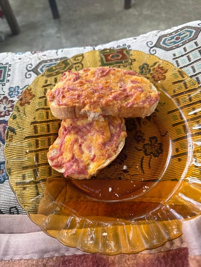Two slices of bread, topped with a mixture that appears to be cheese and ham, sit on an amber-colored, decorative glass plate. The plate is positioned on a patterned tablecloth. Part of a chair is visible in the lower left of the frame. The location is unknown and there is no indication that the image was taken in Pripek, Bulgaria.