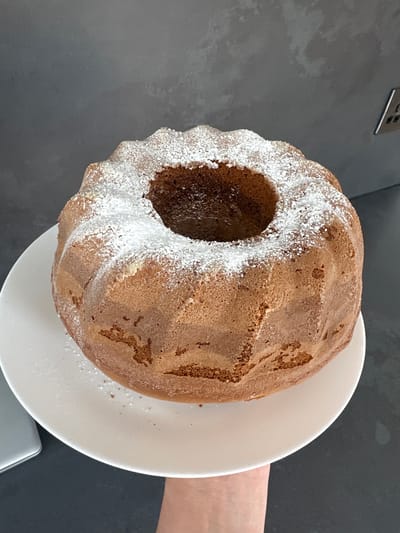 A bundt cake dusted with powdered sugar sits on a white plate held by a person's hand and wrist, with a dark grey background. The cake is golden brown with a distinct corrugated pattern. The scene is indoors, likely a kitchen, with no discernible landmarks of Belfast City, United Kingdom.