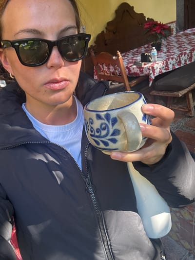 A female subject is presented in a medium-close shot within an outdoor or semi-outdoor patio setting, identified as being in San Juan del Río, Mexico. She wears black Ray-Ban Wayfarer-style sunglasses, a white ribbed crew-neck t-shirt, and a black puffy jacket with a visible zipper and circular pull tab. A small gold-colored hoop earring is visible in her left ear. Her lips are slightly parted. Her right hand holds a light-colored ceramic mug adorned with blue floral-scroll patterns. In the background, to the subject's right, a wooden table is covered with a white tablecloth featuring a red floral design. On the table, a small potted poinsettia plant, a plastic water bottle, and a dark object are visible. Behind the table, a carved wooden chair with a dark cushion and an ornate, dark wooden wall fixture are present. Strong sunlight illuminates the scene, creating distinct shadows. The immediate ground visible is tiled.