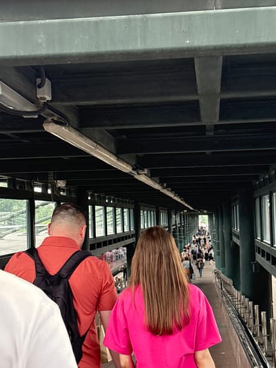The image shows a crowded pedestrian walkway, likely an overpass or elevated bridge, with numerous people walking in both directions. The walkway is enclosed by windows on one side, offering a view of greenery and some structures outside. The ceiling of the walkway is a dark, industrial-looking structure with exposed beams and lighting fixtures.
In the foreground, two individuals are visible from behind. The person on the left is wearing a bright orange short-sleeved shirt and a black backpack. The person on the right is wearing a vibrant pink t-shirt. A portion of a white garment is visible in the lower-left corner, suggesting another person is in front of the orange-shirted individual.
Further down the walkway, a dense crowd of people can be seen moving, many carrying bags or backpacks. The architecture of the walkway appears to be utilitarian, with metal railings along the edge and visible support structures. The lighting suggests it is daytime, possibly overcast, given the diffused light.
The location is identified as the city of Imeni Morozova, Russia, and the scene is consistent with a busy public transit hub or a popular pedestrian thoroughfare within an urban or semi-urban environment. The image captures a moment of transit, with many individuals on their way to various destinations.