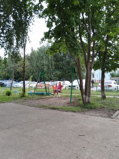 An outdoor, medium-distance, eye-level shot shows a child on a swing set in Roshchinsky, Russia. The child, wearing a pink shirt and shorts, is in motion on a swing. The swing set is green metal with a sand-covered area beneath it. Trees with green leaves are present in the foreground and background. Several cars are parked in the background, and a building is visible behind them. The ground is paved with concrete and grass. The lighting suggests it is daytime.