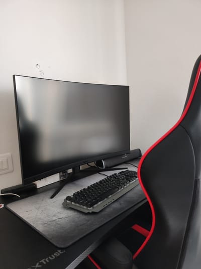 This is a medium shot of a gaming setup in a room. The scene includes a monitor, keyboard, mousepad, a soundbar, and a gaming chair. The monitor is large and curved, displaying a dark, reflective screen. In front of it sits a compact black keyboard with grey keycaps. A grey and black patterned mousepad is beneath the keyboard. A dark grey soundbar is positioned on the monitor stand. A black gaming chair with red trim is visible on the right side of the frame. The setting appears to be indoors, with a plain white wall behind the desk. There are some marks on the wall above the monitor, which could be paint damage or tape residue. A light switch is visible on the wall to the left of the monitor. The overall impression is of a clean and organized gaming station, possibly in a home or office environment. There are no people present. The time of day is indeterminate, but the lighting suggests it might be daytime. No weather is visible. The context provided indicates the location is Grosseto, Italy, but there are no specific visual cues within the image to confirm this. Some text is visible on the desk mat at the bottom of the frame, reading "XTSTrust.".