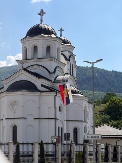 The image shows a white church with black domes, topped with crosses, in Prijepolje Municipality, Serbia. The church is built on a slight incline, with a backdrop of mountains covered in trees. A flag with a cross is waving near the top of the church. A street lamp is visible next to the church. A sign with Cyrillic text, "Сељашница", is attached to a post, and a sign indicating the "Манастир Хуманитарного Попечитељства Богородице" with a picture of the Monastery is also visible. A black iron fence surrounds the perimeter of the church.
