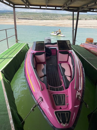 The image depicts a pink speedboat docked on a floating platform covered in artificial turf, with a view of the Chirkey reservoir, located near the city of Chirkey, Russia. The speedboat features a black interior with pink and black seats. A personal watercraft with a person aboard is visible in the distance on the reservoir. A second watercraft rests on the platform behind the speedboat. A covered structure, likely a dock, is visible on the left side of the image. The reservoir shore appears arid with signs of erosion.