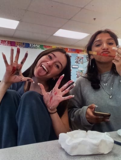 The image displays two young females in an indoor setting, consistent with an art classroom or studio. No specific visual elements within the image identify the location as Aktash, Uzbekistan.
The female on the left has long dark hair and is looking directly at the camera with her tongue extended and a wide smile. Her hands are raised, open, and visibly covered in various colors of paint, including red, white, purple, and black. She wears a ring on her right ring finger and is dressed in a dark-colored top and pants.
The female on the right, also with long dark hair, is looking towards the camera while holding two paintbrushes horizontally across her mouth. She wears a grey sweatshirt and multiple silver necklaces, one featuring a butterfly charm. Her right hand displays several rings, and her left hand holds a dark-colored smartphone.
In the lower right foreground, a textured, crumpled white object, possibly an art project made of clay or papier-mâché, rests on a light-colored, speckled table surface. The background shows a wall with a large bulletin board or whiteboard featuring colored strips and mirrored text, including "EX LINE SPACE," along with a grid of smaller, possibly decorated, squares. Overhead, rectangular fluorescent light panels are visible on the ceiling.