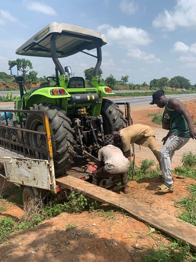 In Sobon-Gida, Nigeria, three men are observed working on a bright green Zoomlion tractor. The tractor is positioned on the back of a white, rustic trailer, which appears to be parked on the side of a road with sparse vegetation and red soil. Two men are bent over, actively engaged with the tractor's rear mechanism, while a third man stands nearby, looking down at them. The environment suggests an outdoor, potentially rural or roadside setting under a bright, cloudy sky.