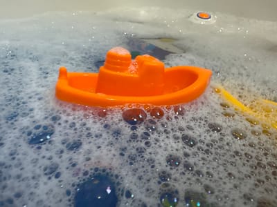 An orange plastic toy boat floats in a bathtub filled with white bubbles and water. The boat is in the foreground and slightly to the left of the image. The bathtub appears to be white with some shiny elements reflecting in the water and bubbles. In the background, there is a small blue object and a hint of a yellow object. The overall scene is a close-up shot of bath toys in soapy water.