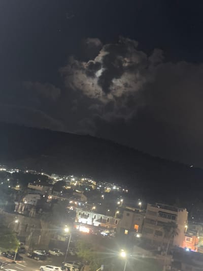 The image depicts a nighttime view of Tiberias, Israel. The sky is dark with some clouds partially obscuring a bright moon. Below the sky is a dark, mountainous silhouette, likely Mount Tiberias. The foreground shows a section of the city with buildings illuminated by streetlights and interior lights. Several cars are visible on the streets, and trees are scattered throughout the urban landscape. The overall impression is of a quiet, residential area in Tiberias at night, with the natural landscape of the surrounding mountains forming a backdrop.