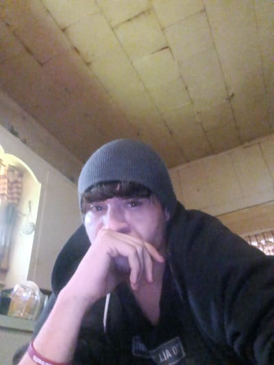 A selfie shows a person wearing a dark grey beanie and a black hooded sweatshirt. The person's hand is raised to their mouth, appearing pensive. A purple bracelet is visible on their left wrist. In the background, there's a ceiling composed of wooden planks, and what appears to be a kitchen area with some items on a shelf. The location is indeterminate.