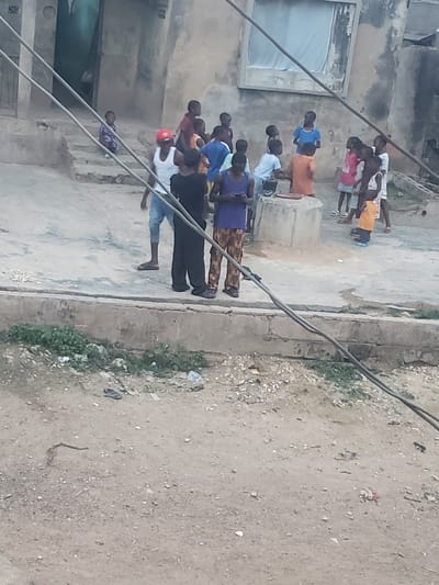 A group of people stand on a street in Alalubosa, Nigeria. The street is bordered by buildings and a dirt area with vegetation. The people appear to be observing something, possibly performing an action. Power lines are visible in the foreground. Some litter is scattered on the ground.